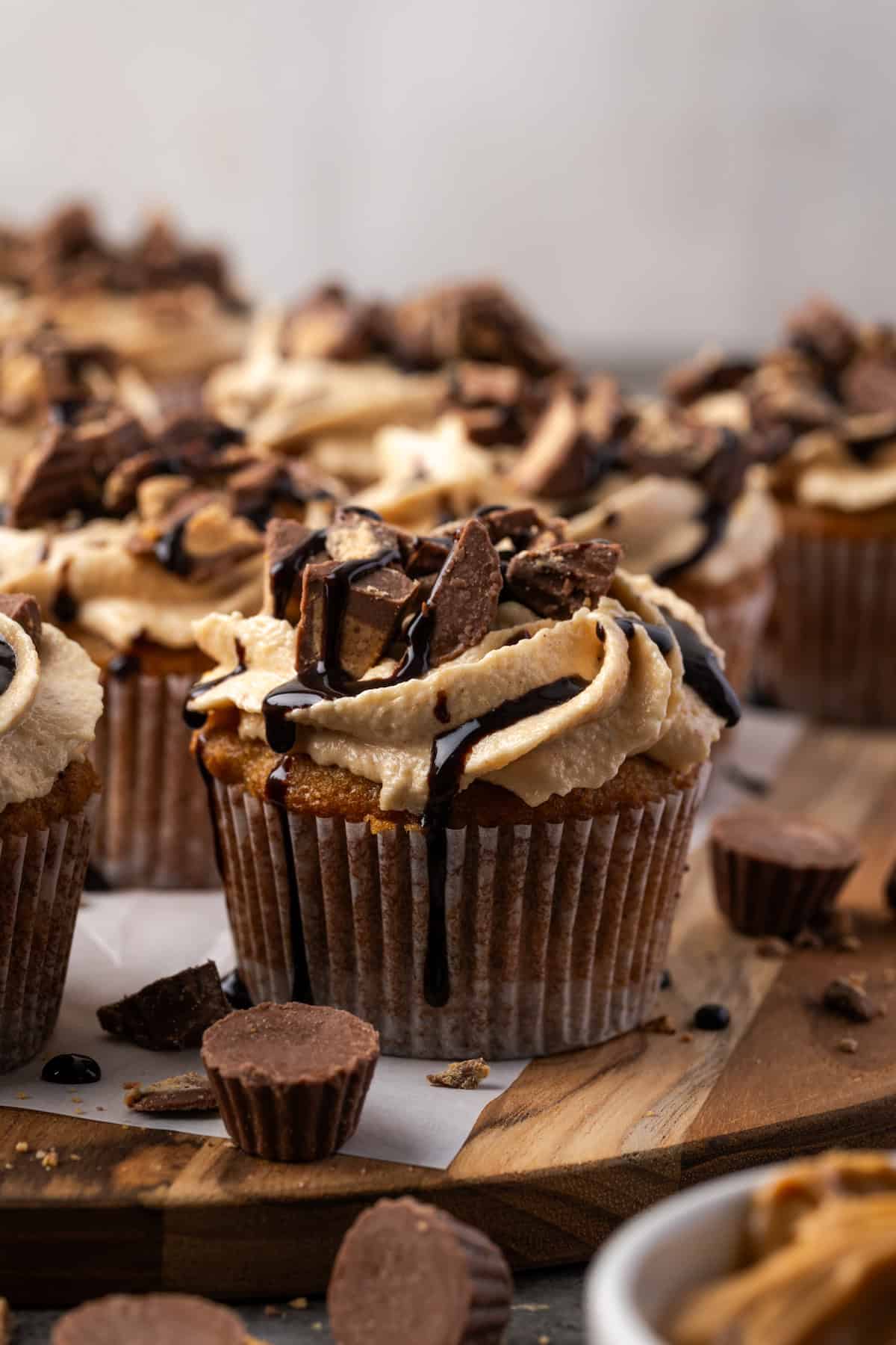 A frosted banana cupcake drizzled with chocolate, with more cupcakes in the background and peanut butter cups scattered in the foreground.