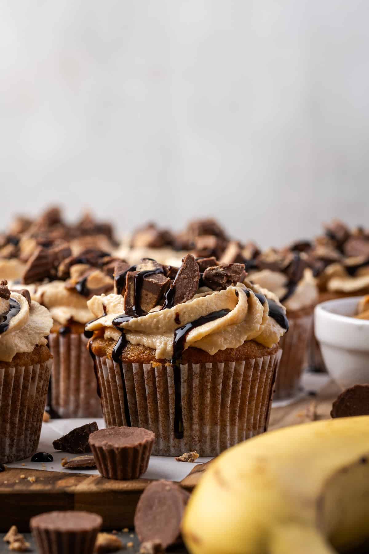 Assorted frosted banana cupcakes drizzled with chocolate, with a banana and peanut butter cups in the foreground.