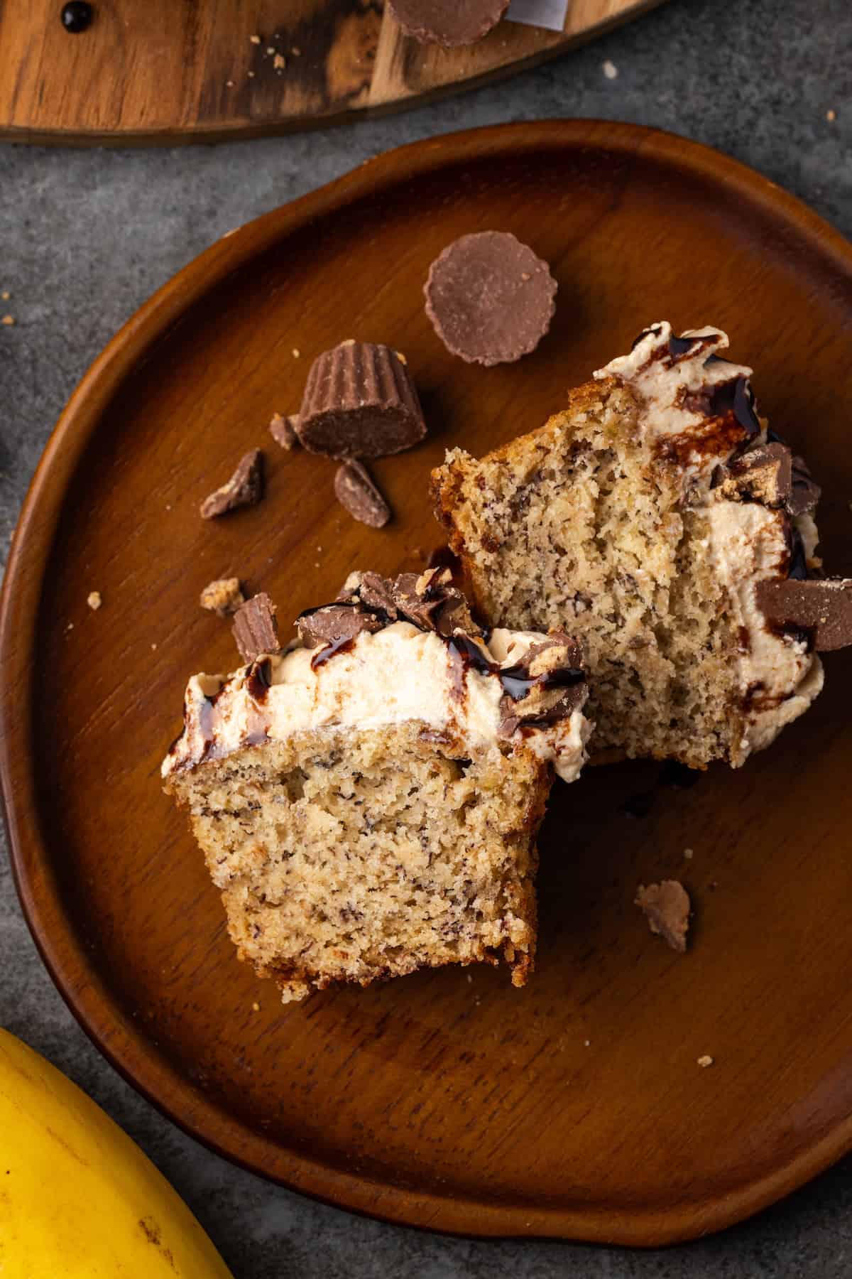 Overhead view of two halves of a frosted banana cupcake next to chopped peanut butter cups on a brown stoneware plate.