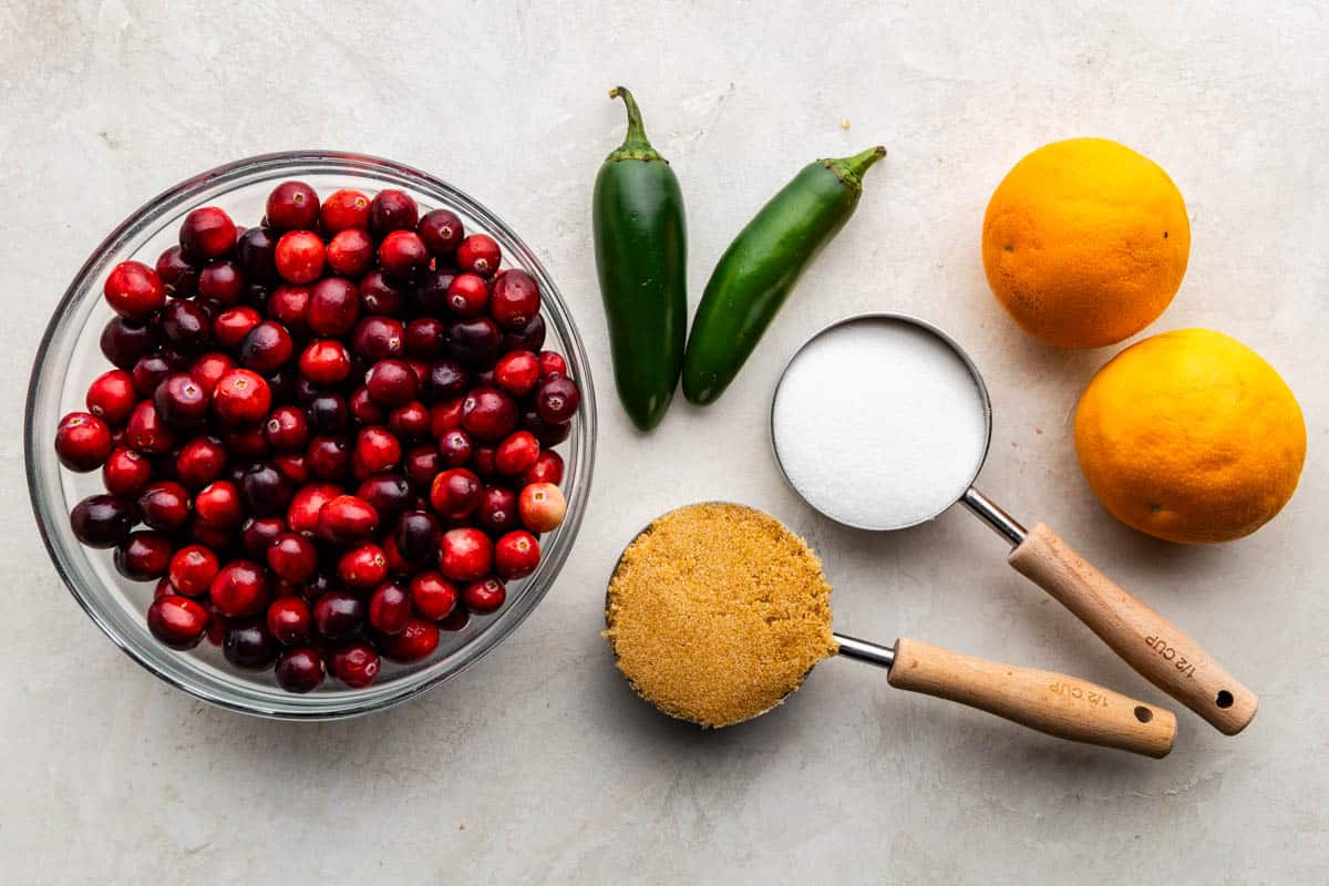 An overhead view of a bowl filled with fresh cranberries, set next to other raw, wet, and dry ingredients needed to make jalapeno cranberry sauce.