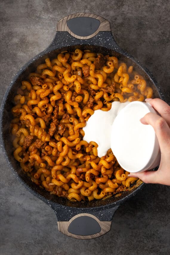 Milk and sour cream mixture being poured over the cheeseburger noodle mixture in a skillet.