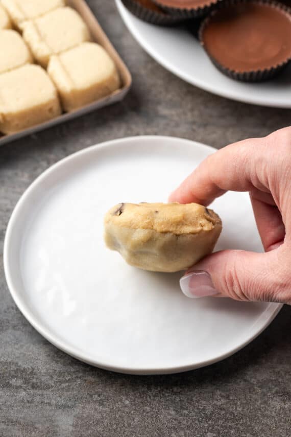 A hand holding an unbaked cookie cup made from chocolate chip cookie dough and sugar cookie dough wrapped around a Reese's peanut butter cup, with a plate below.