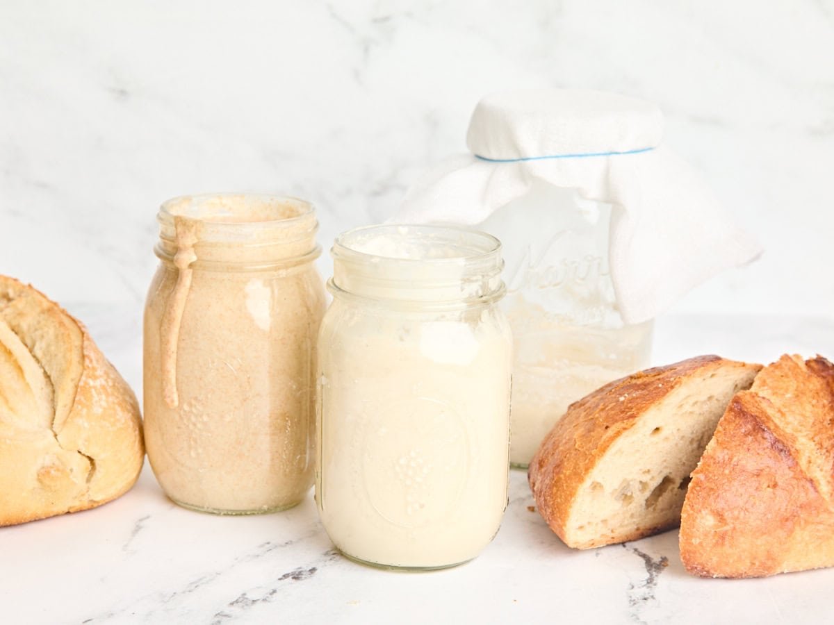 Side view of two jars of homemade sourdough starter with a loaf of sourdough bread.